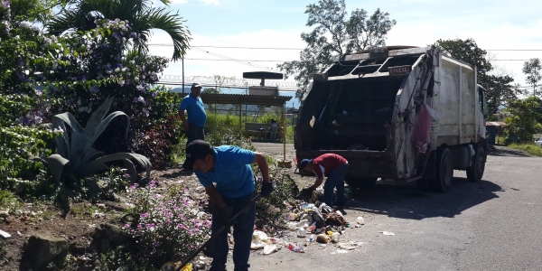 Trabajadores con camisas azules y rojas atendiendo tareas comunitarias junto a un camión, día soleado con plantas floridas
